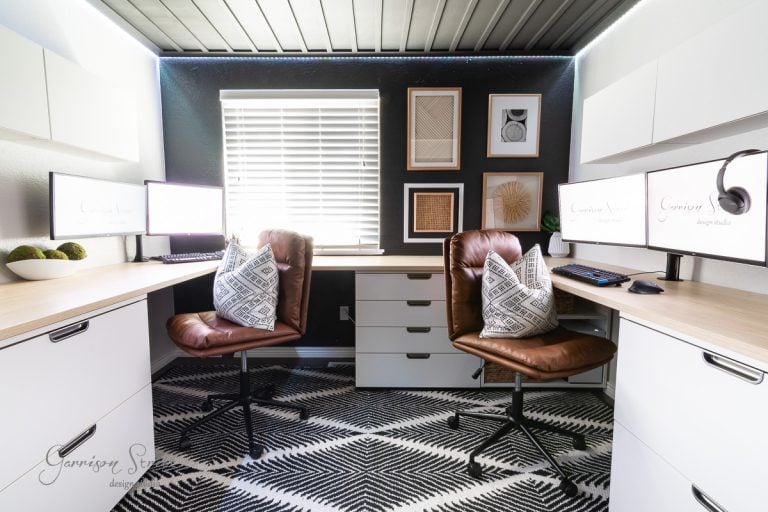 A modern home office featuring a diy wood plank ceiling, with two brown leather chairs, a black and white geometric rug, dual monitors on L-shaped desks, and decorative wall art. A window with blinds lets in natural light.