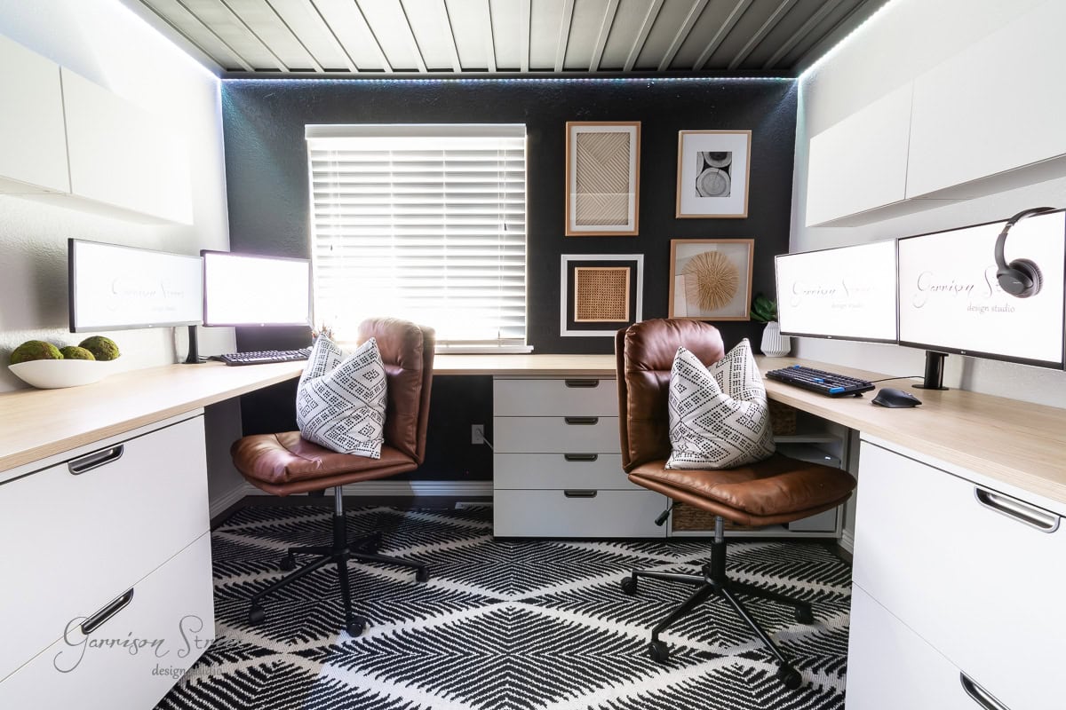 A modern home office featuring a diy wood plank ceiling, with two brown leather chairs, a black and white geometric rug, dual monitors on L-shaped desks, and decorative wall art. A window with blinds lets in natural light.