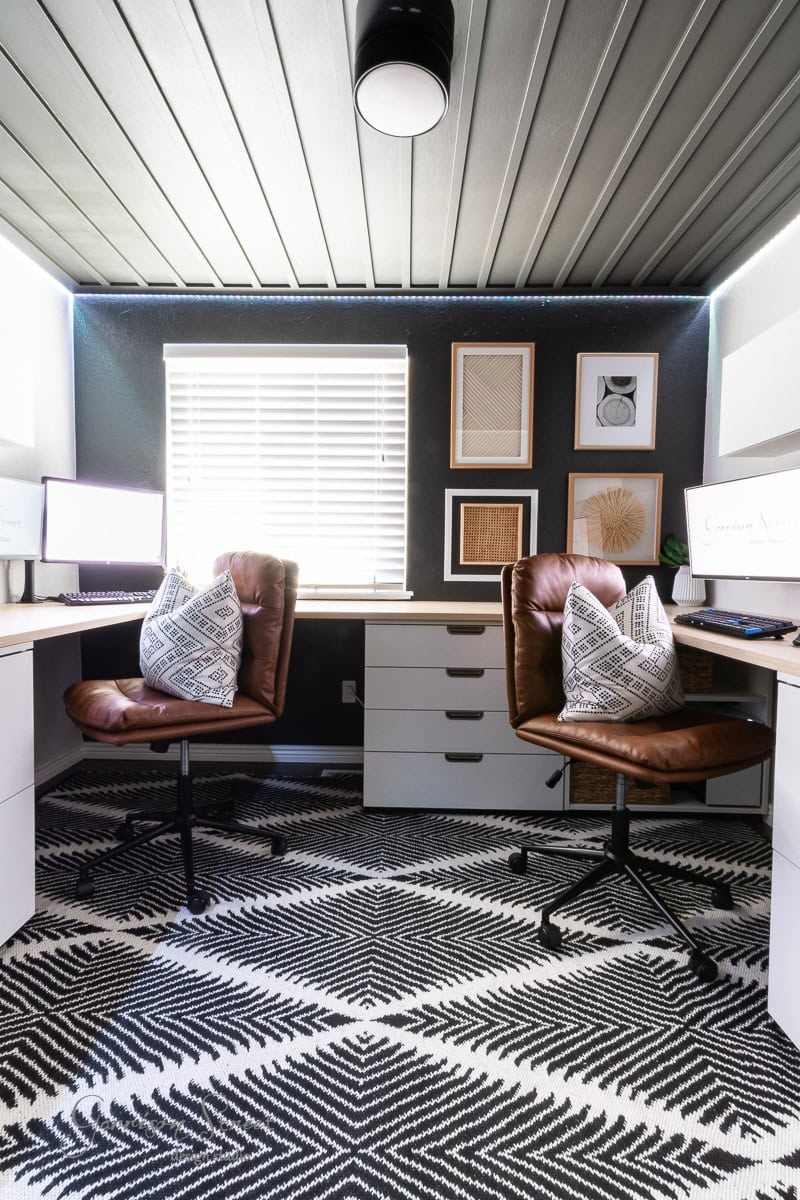 A modern office space with two brown leather chairs, each with a patterned pillow. Desks with computer monitors flank the room, and a geometric black-and-white rug covers the floor. Above it all, a DIY wood plank ceiling adds warmth. Three framed wall art pieces hang above. Light filters through blinds.
