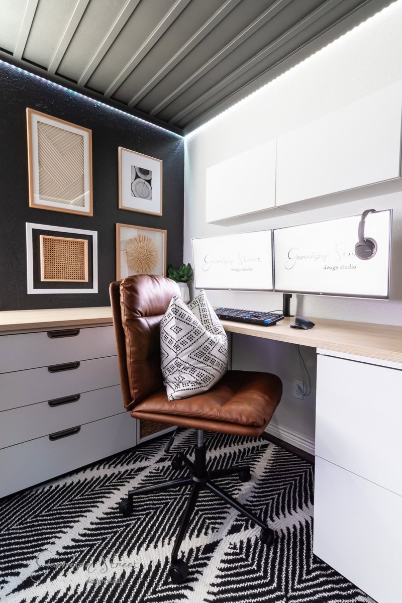 A modern home office with a sleek desk featuring dual monitors and a brown leather chair with a patterned pillow. The DIY wood plank ceiling adds warmth, while the wall displays abstract artwork and a geometric black-and-white rug covers the floor.