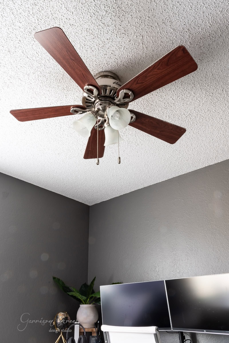 Ceiling fan with five wooden blades and three light fixtures, perfectly complementing the DIY wood plank ceiling. Below, a corner of the room showcases a desk, computer monitors, and a potted plant, enhancing the cozy yet modern workspace vibe.