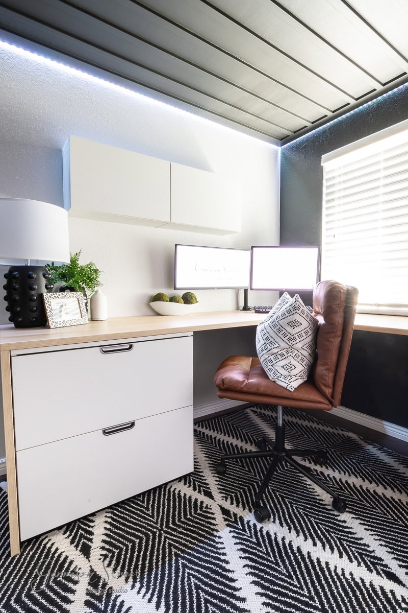A modern home office with a white corner desk and dual monitors sits beneath a stylish DIY wood plank ceiling. A brown leather chair is in front of the desk, while decorative plants and a pillow add flair. A geometric patterned rug covers the floor as natural light streams through the window.