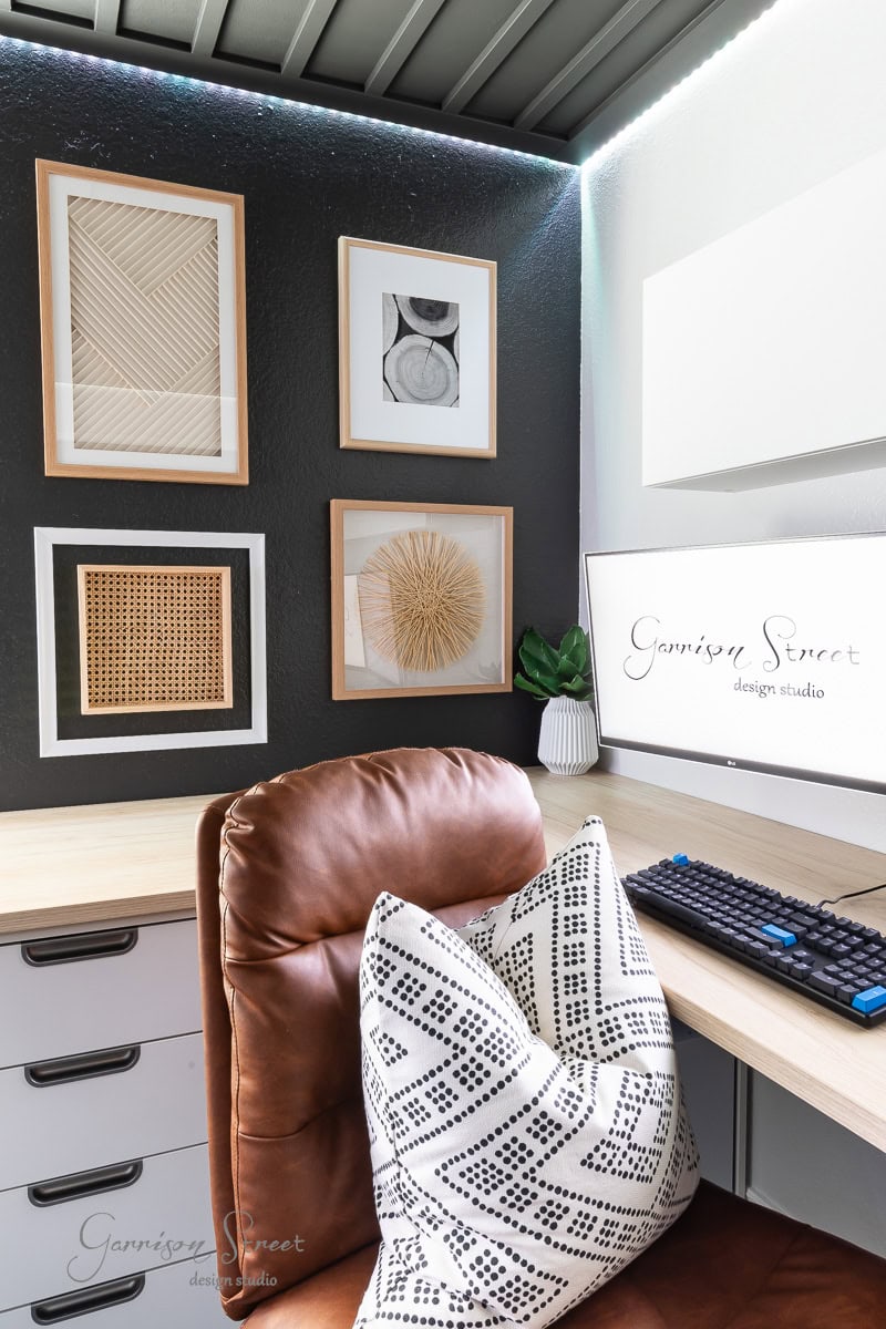 A modern home office corner features a brown leather chair and a black-and-white patterned pillow. The desk holds a keyboard and monitor displaying "Garrison Street Design Studio." Geometric wall art, a DIY wood plank ceiling, and a small plant enhance the room's dark tones.