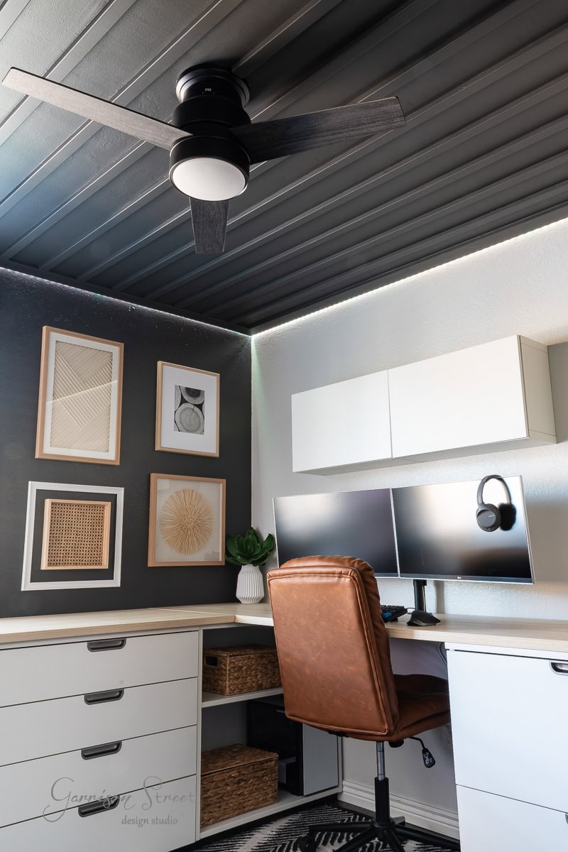 A modern home office features a brown leather chair, two computer monitors on a white desk, and a DIY wood plank ceiling with a sleek fan. A white wall cabinet and framed abstract art decorate the space for a minimalist look.