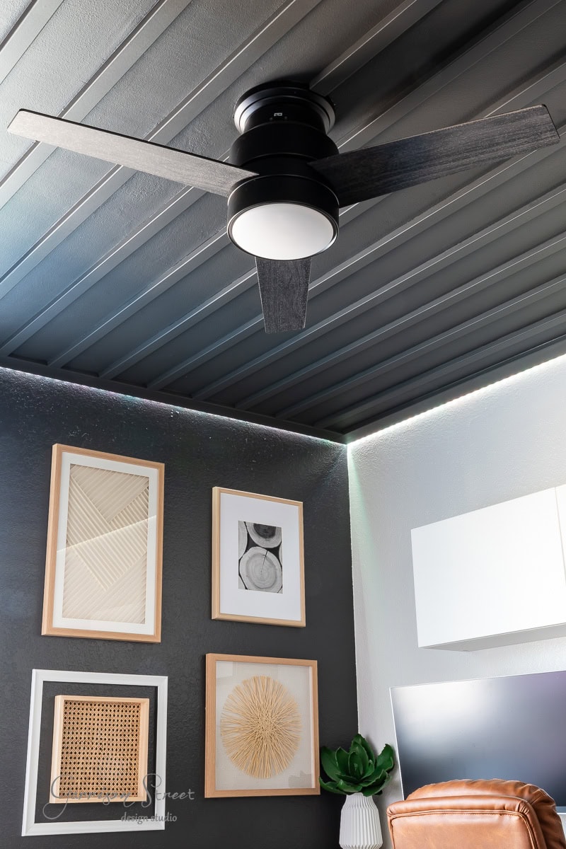 A modern room features a DIY wood plank ceiling, enhancing its elegance. A black ceiling fan complements four framed abstract art pieces on a dark wall. A plant and part of a computer monitor are visible in the corner, adding life and functionality to the space.