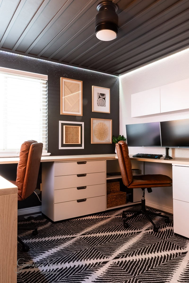 A modern home office featuring a stunning DIY wood plank ceiling, two desks with leather chairs, dual monitors, and a black and white patterned rug. The walls boast framed geometric art, while a window with blinds invites natural light to fill the room.