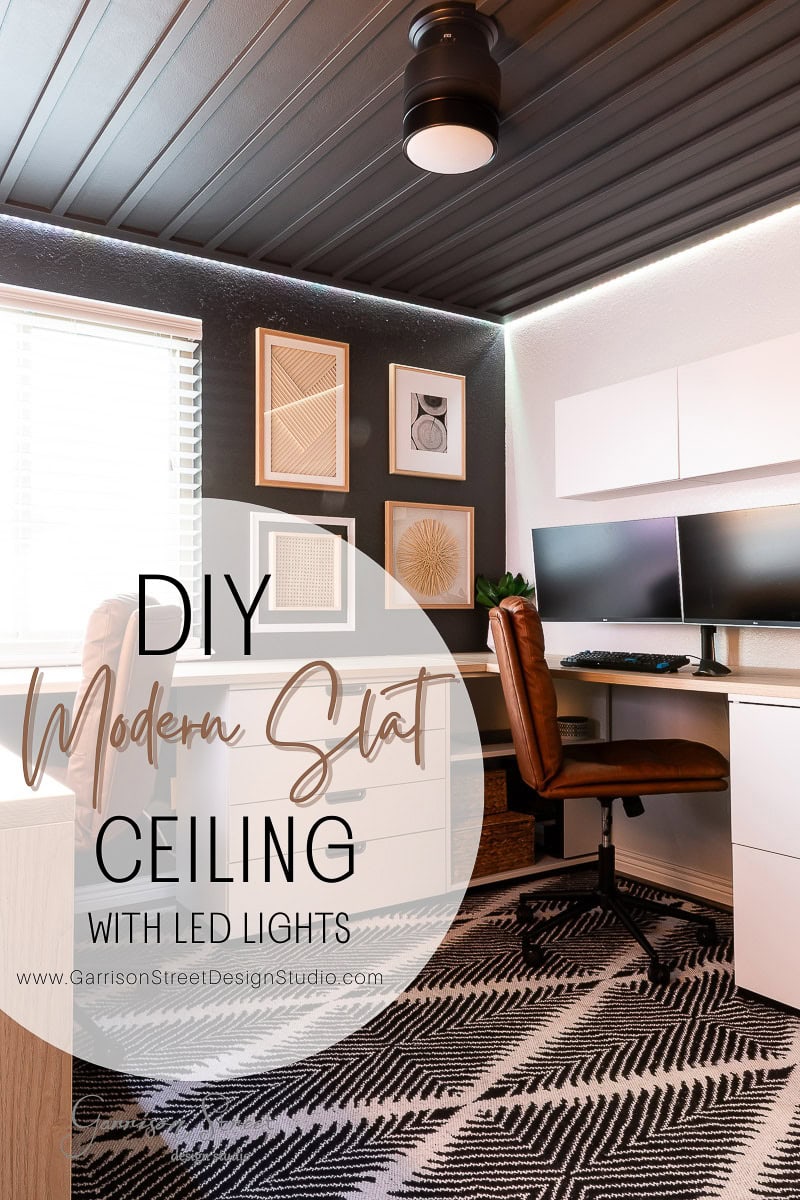 Modern office with a sleek DIY wood plank ceiling and LED lights. Features a brown chair at a white desk with dual monitors. Art decorates the wall, and a patterned rug covers the floor. Text overlay reads: "DIY Modern Slat Ceiling with LED Lights.