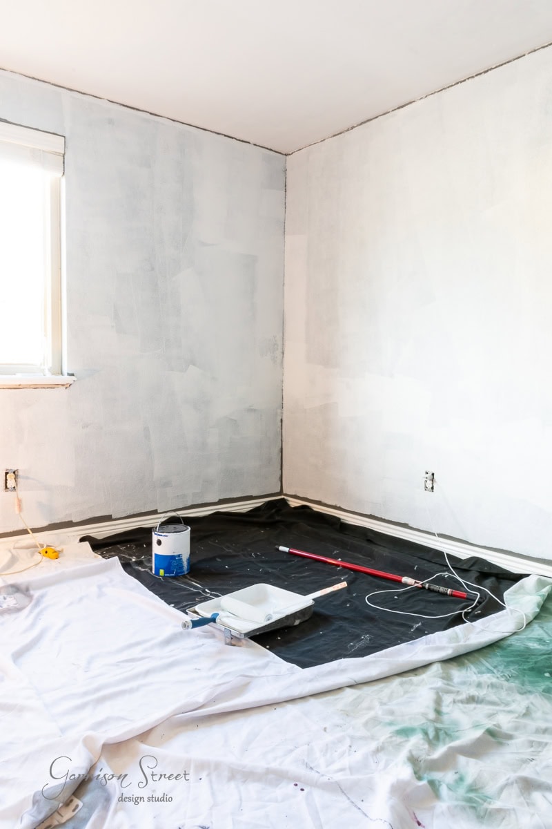 A room corner with freshly painted light gray walls and a DIY wood plank ceiling. A paint can and roller tray with tools are on a protective sheet covering the floor. A window on the left lets in natural light.