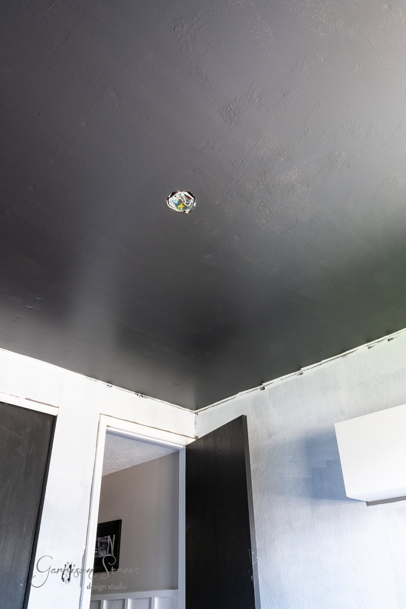 A room with a black ceiling featuring a DIY wood plank design and partially painted white walls. An electrical fixture is visible in the ceiling. The space also includes a black door and a sleek white cabinet.