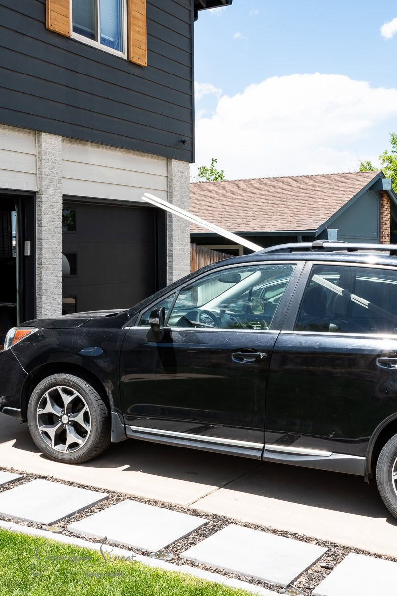 A black SUV is parked in a driveway with long wooden planks protruding from a rear window, likely for a DIY wood plank ceiling project. They extend towards a residential garage under the clear, sunny sky, while a well-manicured lawn flanks the driveway.