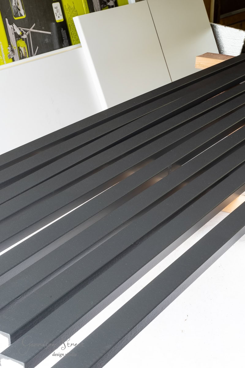 A row of evenly spaced, black metal slats laid out on a surface, supported by wooden blocks that echo the aesthetic of a DIY wood plank ceiling. The background features white panels and product packaging leaning against a wall.