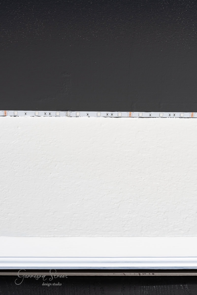 A wall is divided into two sections: the top half is painted black, and the bottom half white. A strip of LED lights marks where they meet, casting a soft glow beneath a DIY wood plank ceiling. A white baseboard runs along the bottom.