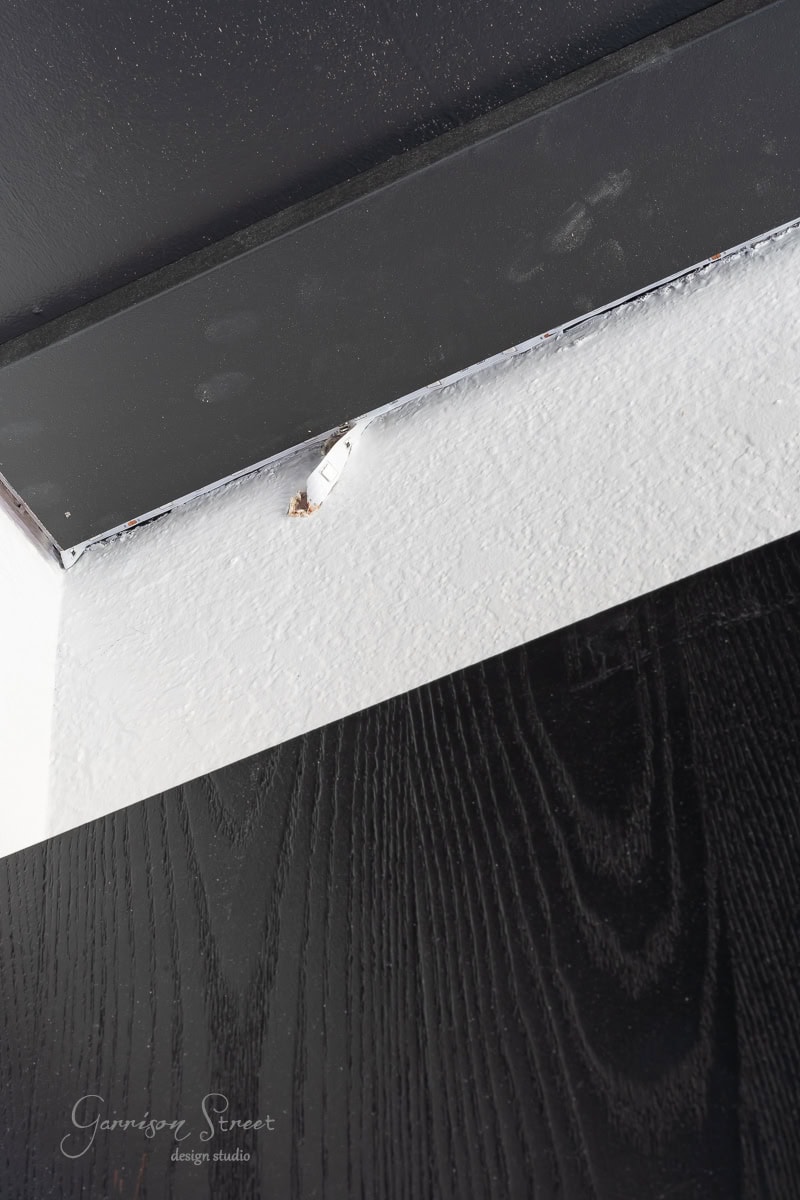 Close-up of a small hole in a white ceiling corner, near a black-painted section, suggesting a past diy wood plank ceiling project. A bit of debris is visible at the edge of the hole.
