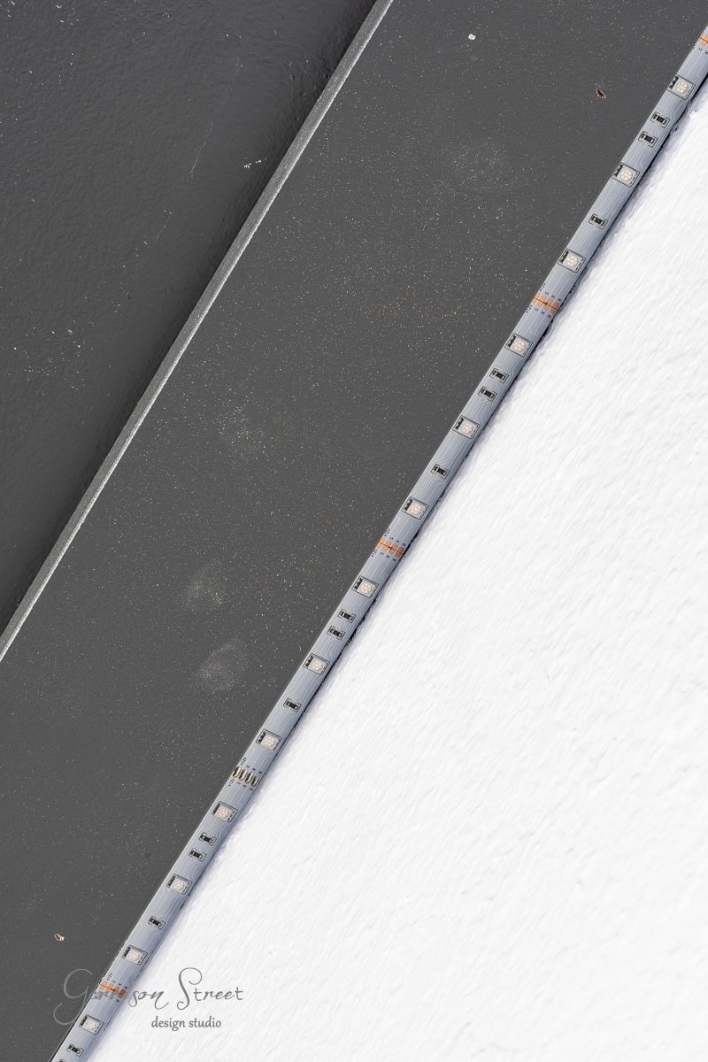 A strip of LED lights is affixed to the edge where a dark gray surface meets a white ceiling, possibly enhancing a DIY wood plank project. The LED strip is positioned diagonally, with faint logo text visible on the bottom left corner.