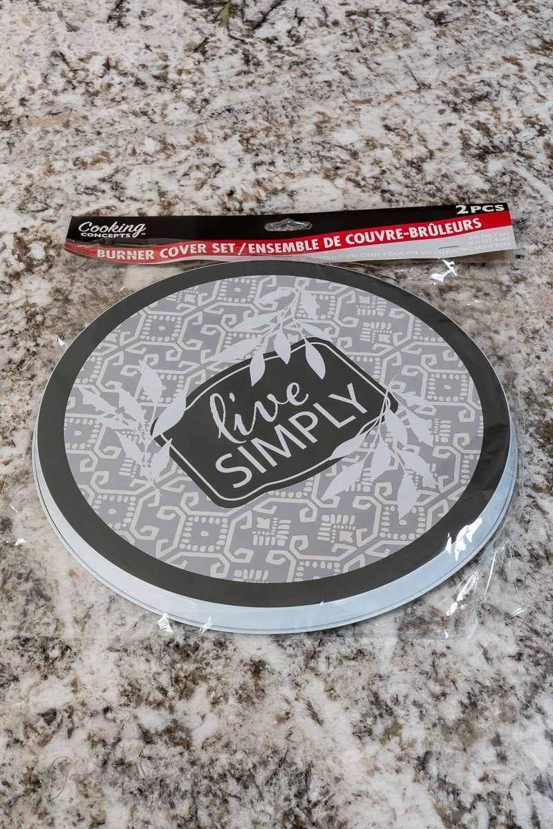 A round burner cover set placed on a granite countertop. The cover, perfect for any outdoor DIY project, features a gray pattern with the phrase "live SIMPLY" in the center, surrounded by decorative leaves. It is packaged with a label that includes the text "Burner Cover Set.