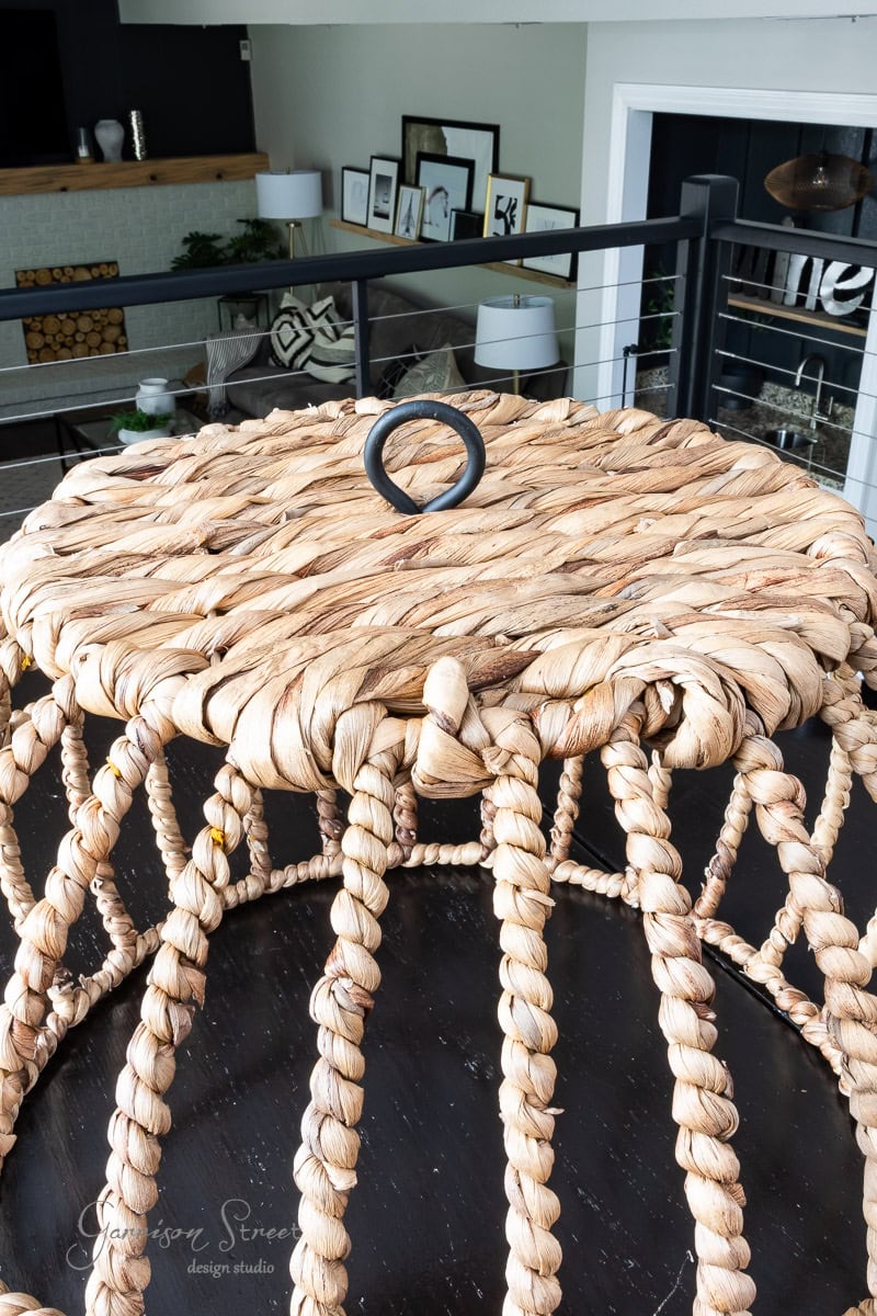A woven rattan side table with a looped handle on top sits on a dark surface, perfect for showcasing your DIY creativity. In the background, a modern living room with framed artwork and decorative accents is partially visible.