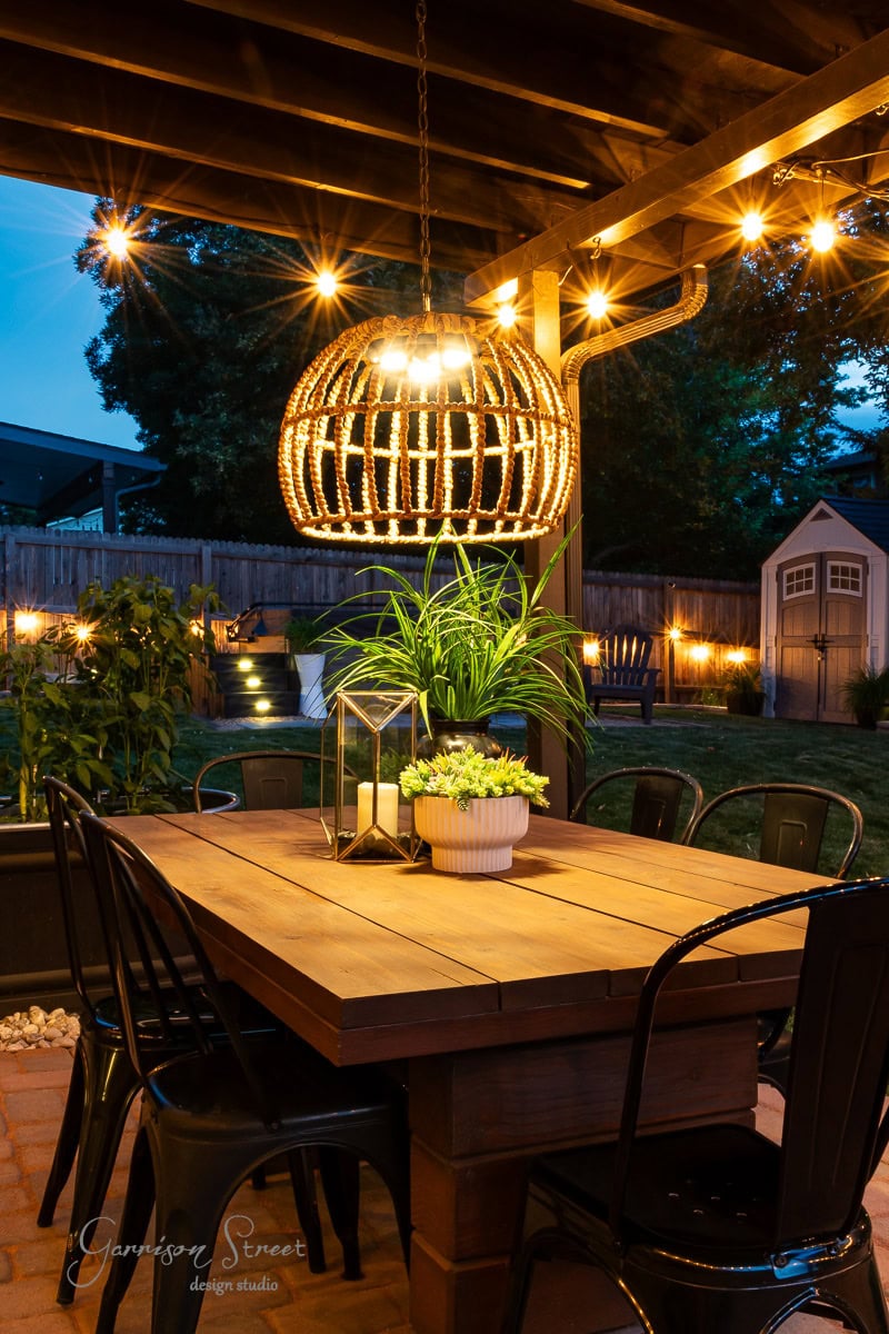 The outdoor patio area at night features a wooden dining table and black chairs, illuminated by a large wicker pendant lamp and string lights. A DIY outdoor chandelier adds charm, while potted plants and a lantern centerpiece adorn the table, with a garden shed visible in the background.