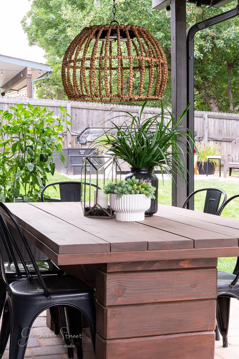 A rustic outdoor patio features a wooden table with metal chairs, topped with green potted plants. A DIY wicker pendant lamp hangs above like an elegant outdoor chandelier. The background shows a wooden fence and greenery, creating a cozy and inviting atmosphere.