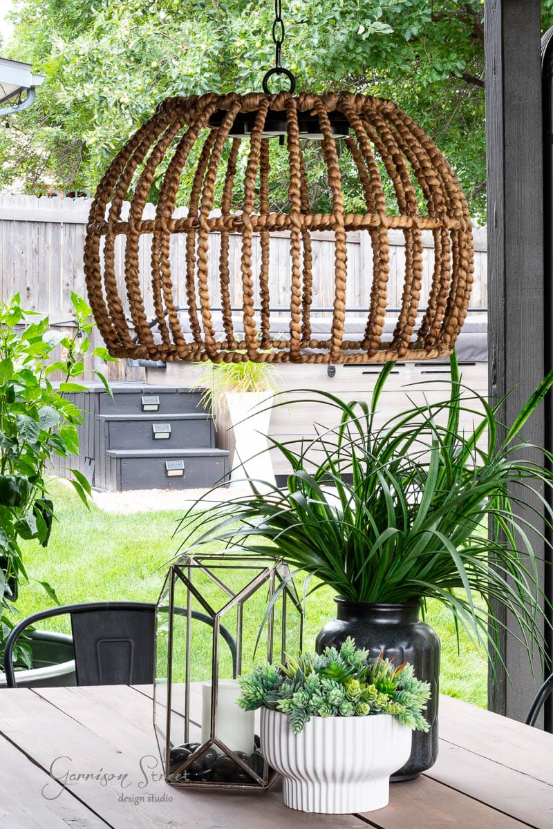 A cozy outdoor patio features a DIY woven rattan pendant light resembling an elegant chandelier hanging over a wooden table. The table is adorned with potted plants, including succulents and greenery, in decorative containers. In the background, a lush garden completes the tranquil scene.