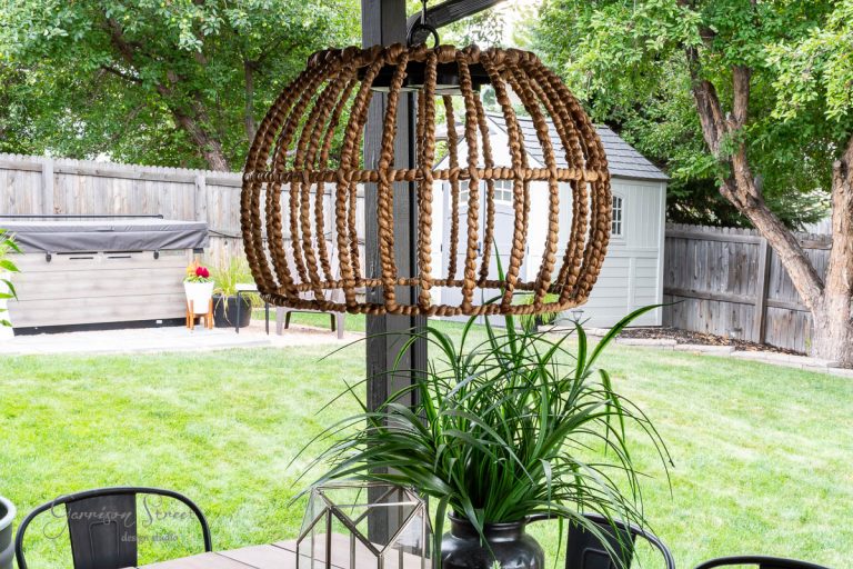 A wooden patio with outdoor furniture features a striking outdoor chandelier above the table. A potted plant adds greenery, and a barbecue grill is seen in the background against the wooden fence. A shed and trees complete this charming DIY-inspired yard scene.