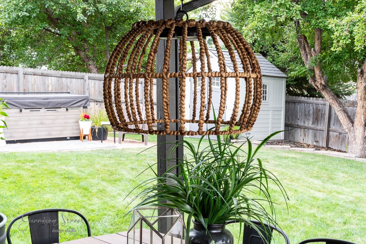 A wooden patio with outdoor furniture features a striking outdoor chandelier above the table. A potted plant adds greenery, and a barbecue grill is seen in the background against the wooden fence. A shed and trees complete this charming DIY-inspired yard scene.