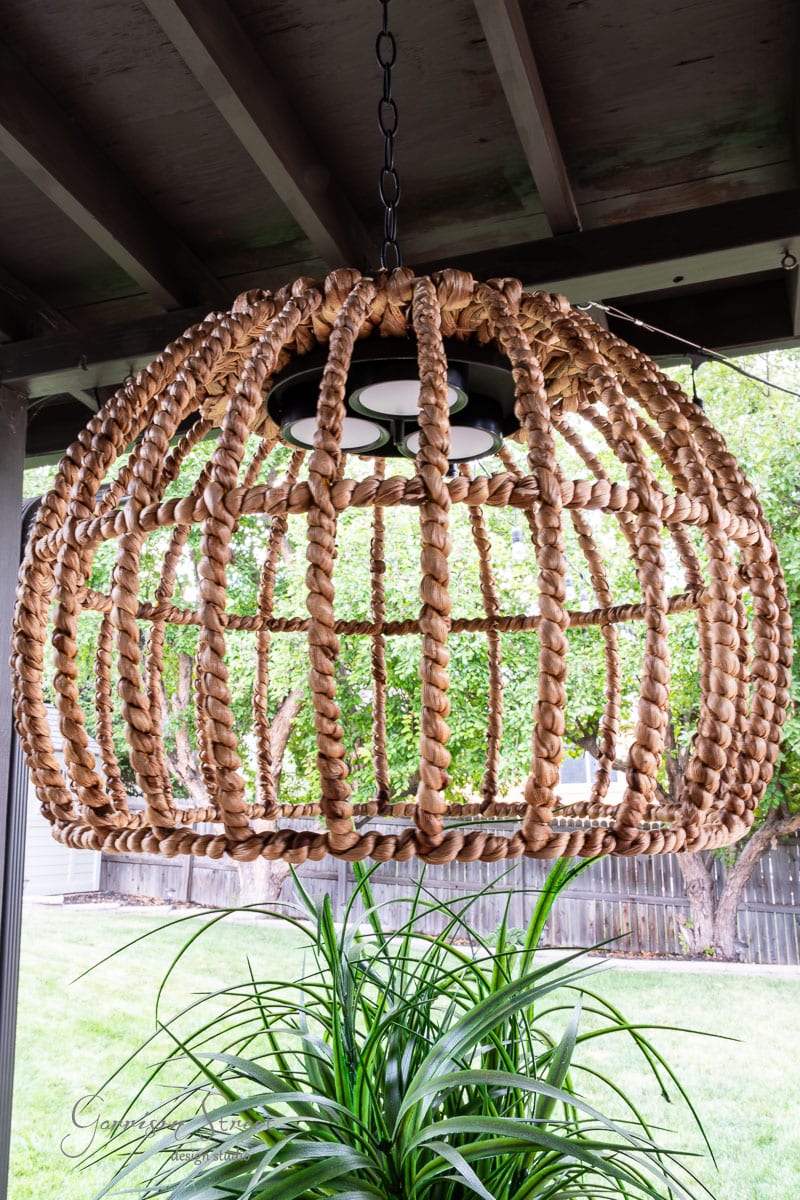 A large, dome-shaped woven rattan light fixture, reminiscent of a DIY outdoor chandelier, hangs elegantly from the ceiling. Below it, a lush green plant thrives against the backdrop of a wooden fence and trees.