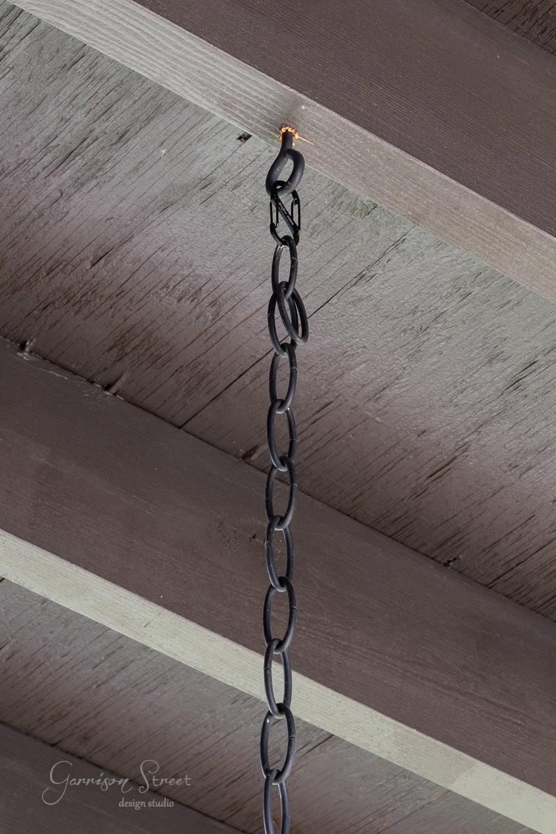 A close-up of a black chain hanging from a wooden ceiling beam, attached by a carabiner. The background shows the textured wood grain reminiscent of an outdoor chandelier project. The words "Garrison Street Design Studio" are visible in the corner.