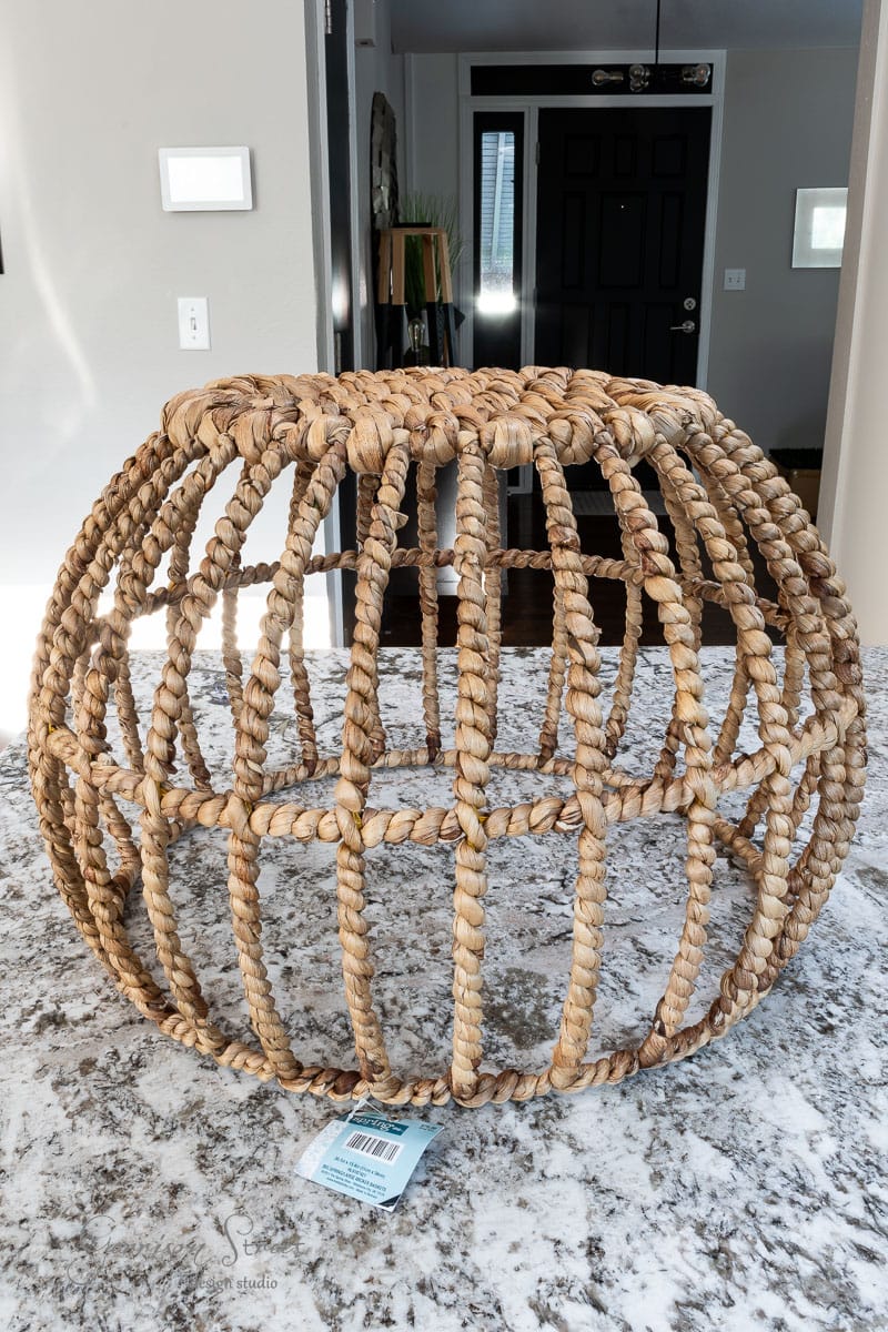 A large, spherical woven basket made of natural materials sits on a marble countertop, reminiscent of an outdoor chandelier. The open weave design creates a lattice structure. The background shows a modern interior with a door, wall trim, and some greenery.