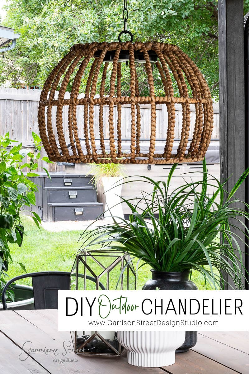 A cozy outdoor space showcases an outdoor chandelier DIY project with a wicker fixture hanging above the table. A potted plant graces the tabletop, and a backdrop of wooden fence and greenery adds serenity. Below, a sign proudly reads "DIY Outdoor Chandelier.