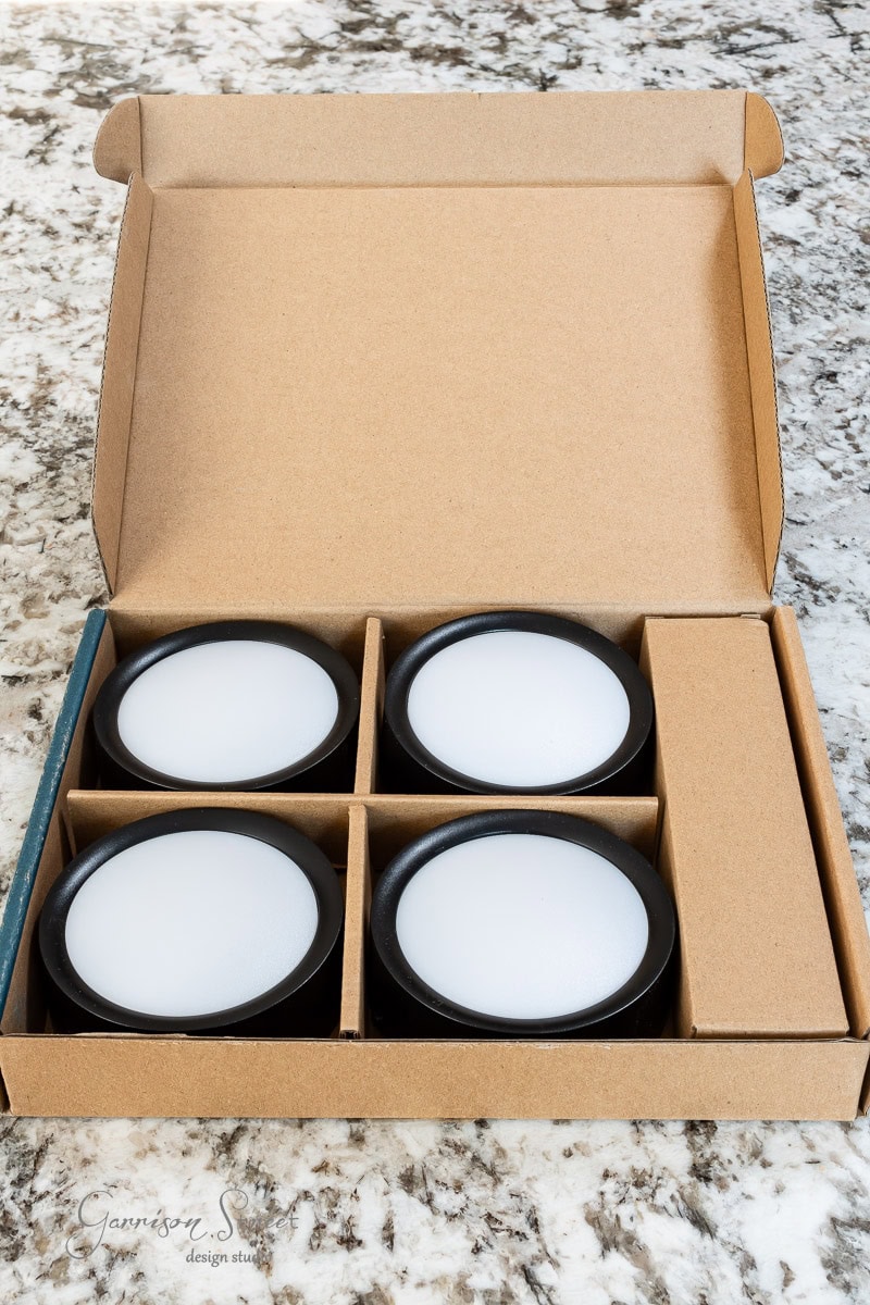 Open cardboard box on a marble surface with four round black and white LED puck lights neatly arranged inside, perfect for your DIY outdoor chandelier project.