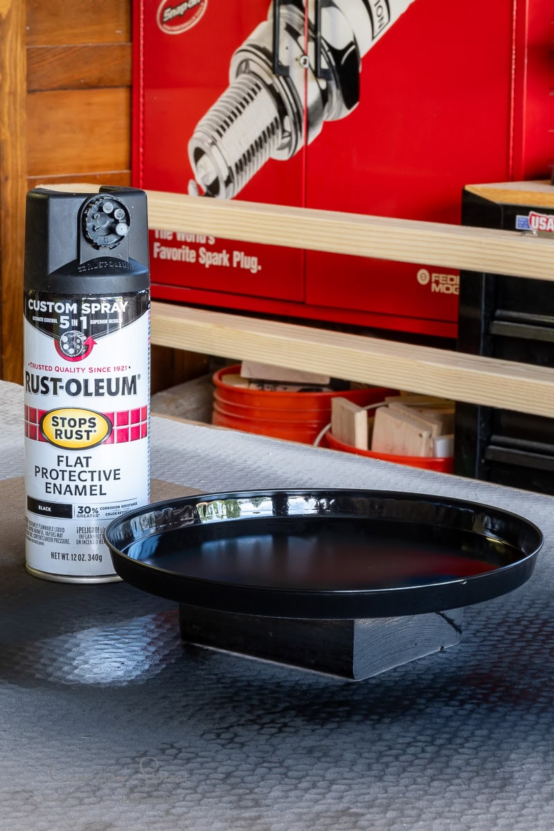 A can of Rust-Oleum flat protective enamel spray paint sits beside a freshly painted black circular metal tray, intended for an outdoor chandelier DIY project, resting on a small wooden stand. Wooden shelves and a wall adorned with a spark plug poster complete the backdrop.