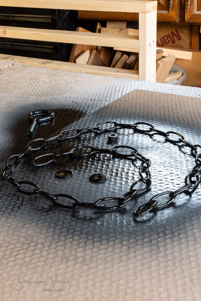 A black chain and hook are laid out on a textured, metallic surface, hinting at a DIY project in progress. Various wooden pieces and cardboard boxes linger in the background, evoking a workshop or studio setting perfect for crafting an outdoor chandelier.