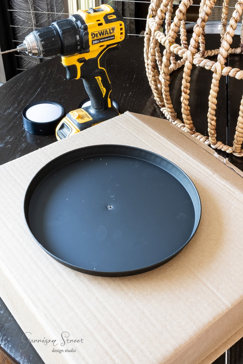 A cordless DeWalt drill with a battery pack sits on a wooden table, ready for your next DIY project. Nearby, a large round black tray and a cardboard box await use, while a woven decorative item serves as inspiration for your outdoor chandelier creation in the background.