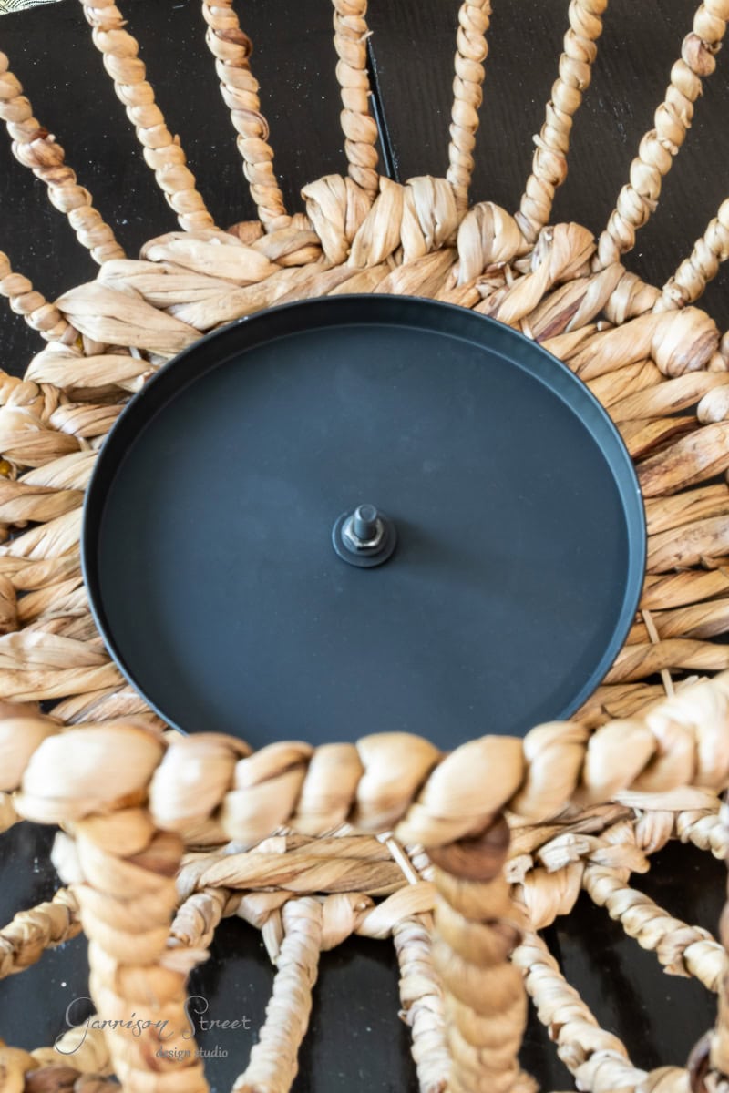 Close-up of a woven wicker light fixture, resembling an outdoor chandelier, with a central black metal plate and socket set against a dark background. The interwoven natural fibers create a radial pattern around the circular metal component.