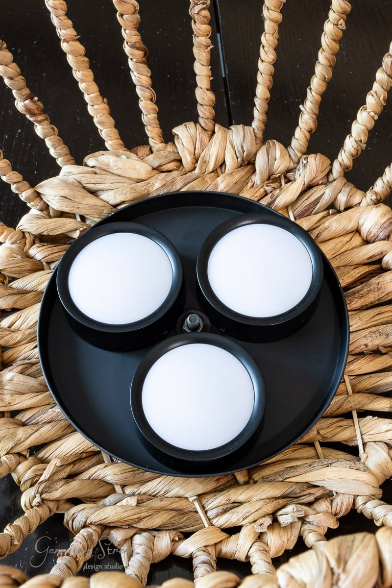 A black tray with three round, white candles sits elegantly on a woven, natural-fiber chair. The minimalist candles contrast with the intricate texture of the chair, creating an ambiance reminiscent of an outdoor chandelier's DIY charm.