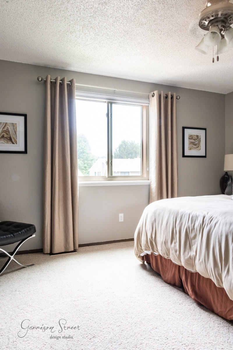 A large window adorned with beige curtains. The room showcases a bed with a white comforter and orange bed skirt, a stylish black bench, and framed artwork on the gray walls. Above, a ceiling fan with a light completes the room.