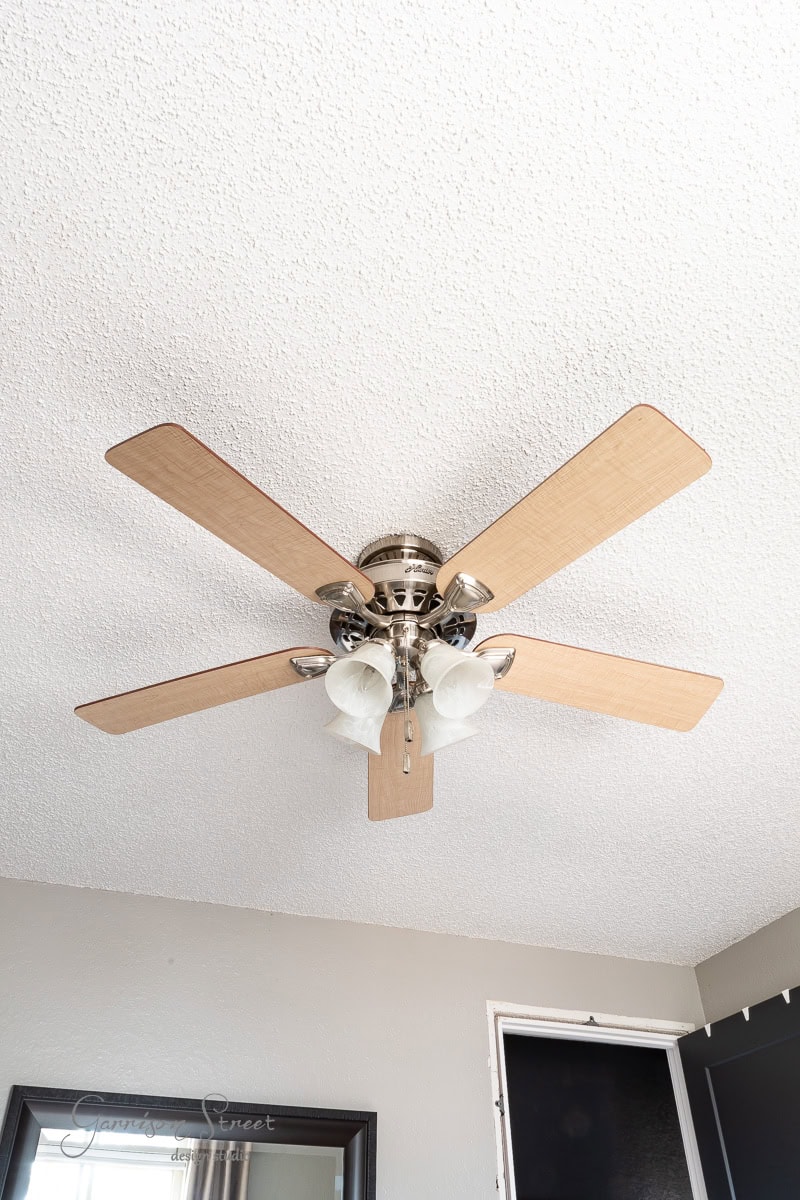 A ceiling fan with five wooden blades and three glass light fixtures hangs from a textured white ceiling. The room includes part of a mirror and a partially visible door.