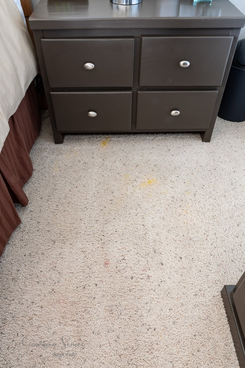 A dark brown dresser with four drawers sits on beige carpeted flooring. The carpet has visible stains, one yellow and one reddish. Part of a bed with a brown bed skirt is on the left side of the image.