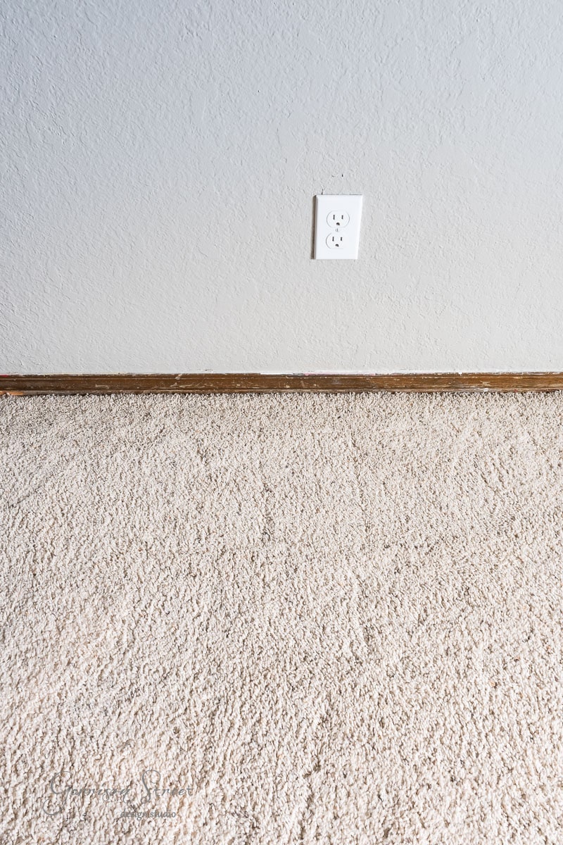 Carpeted floor with beige texture beneath a plain white wall featuring a single electrical outlet. A simple wooden baseboard runs along the wall at floor level.