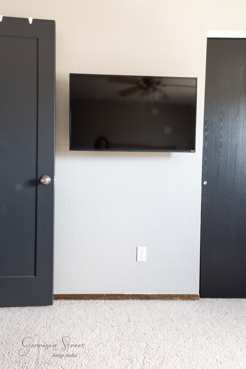 A flat-screen TV is mounted on a beige wall between two dark-colored doors. Below the TV is a white electrical outlet. The light-colored carpet complements the scene, while a ceiling fan subtly reflects on the screen.