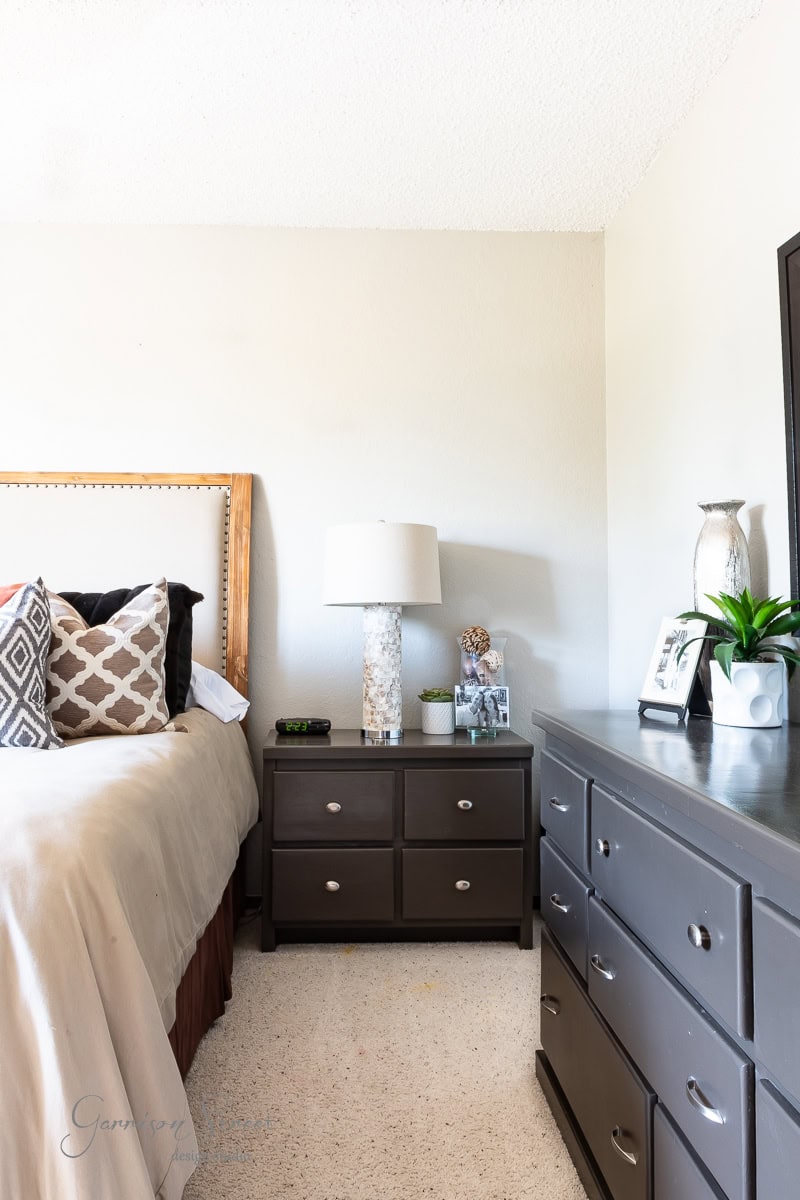 A bed with white and brown pillows, a wooden headboard, and a beige comforter. A nightstand with a lamp and decorative items sits beside the bed, while a dark dresser adorned with a plant and decor enhances the room.