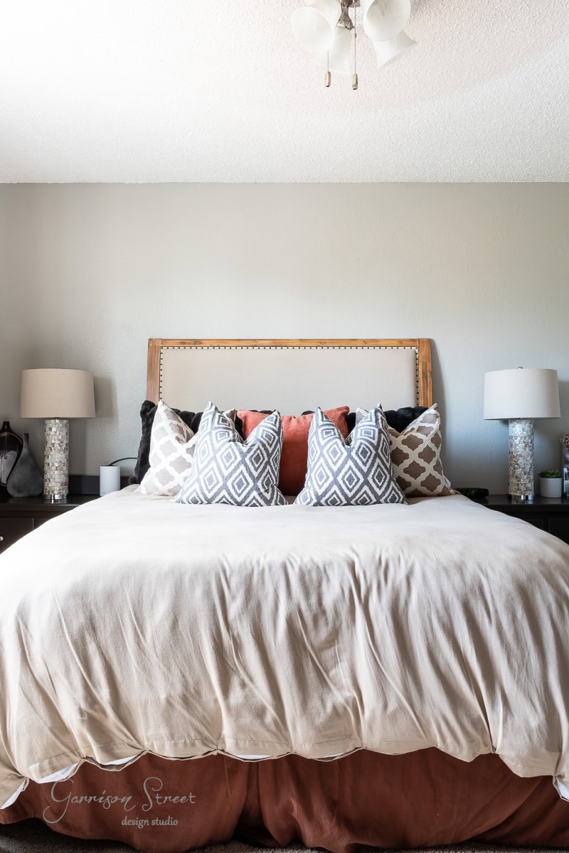 A neatly made bed with a beige and brown comforter is adorned with patterned and solid throw pillows. The simple, elegant headboard complements two bedside tables holding matching lamps with textured bases against a pale gray wall.
