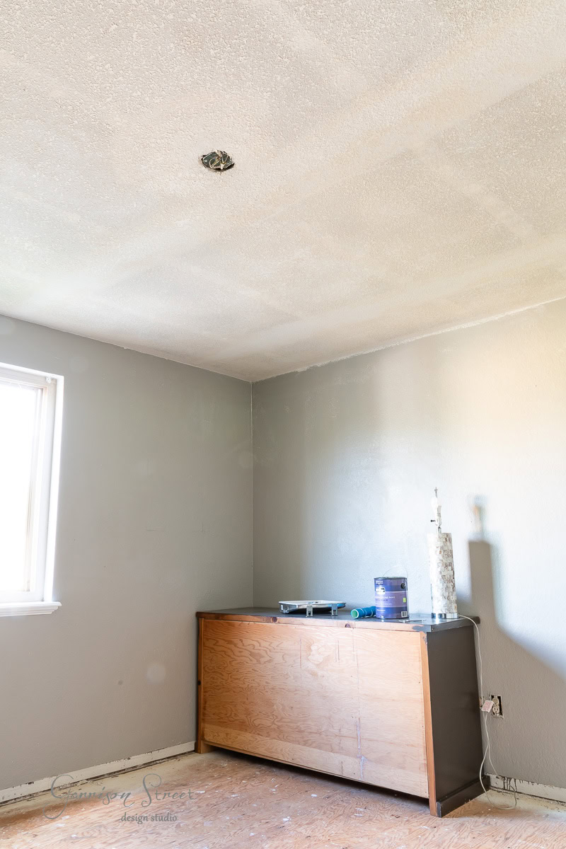 An unfinished room with light gray walls and a white ceiling features a window that lets in natural light. Against the wall, a wooden cabinet holds a small lamp and some paint supplies. The bare floor awaits finishing touches.