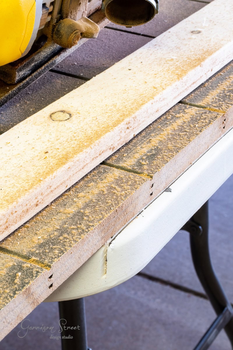 A close-up of a wood plank with sawdust on a table. A yellow circular saw is partially visible at the top. The plank shows multiple cut marks, and the textured table surface embodies craftsmanship in progress.