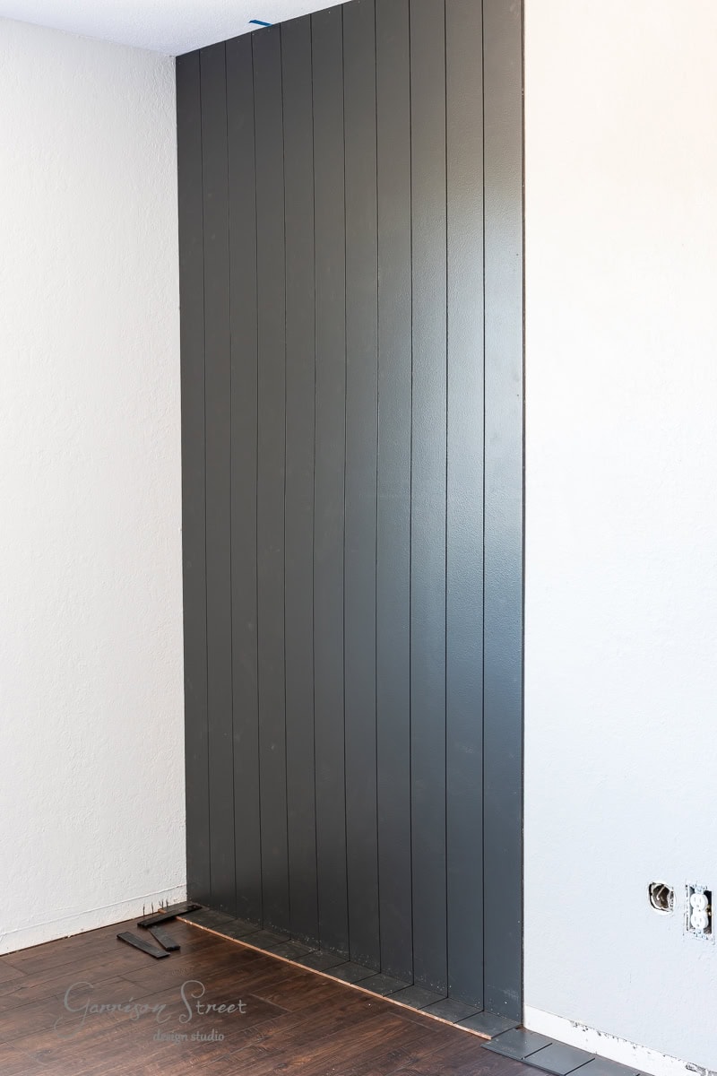 Dark vertical wall panels form a striking accent wall against a white backdrop and wooden floor. Electrical outlets punctuate the design, while unfinished edges hint at ongoing renovations.
