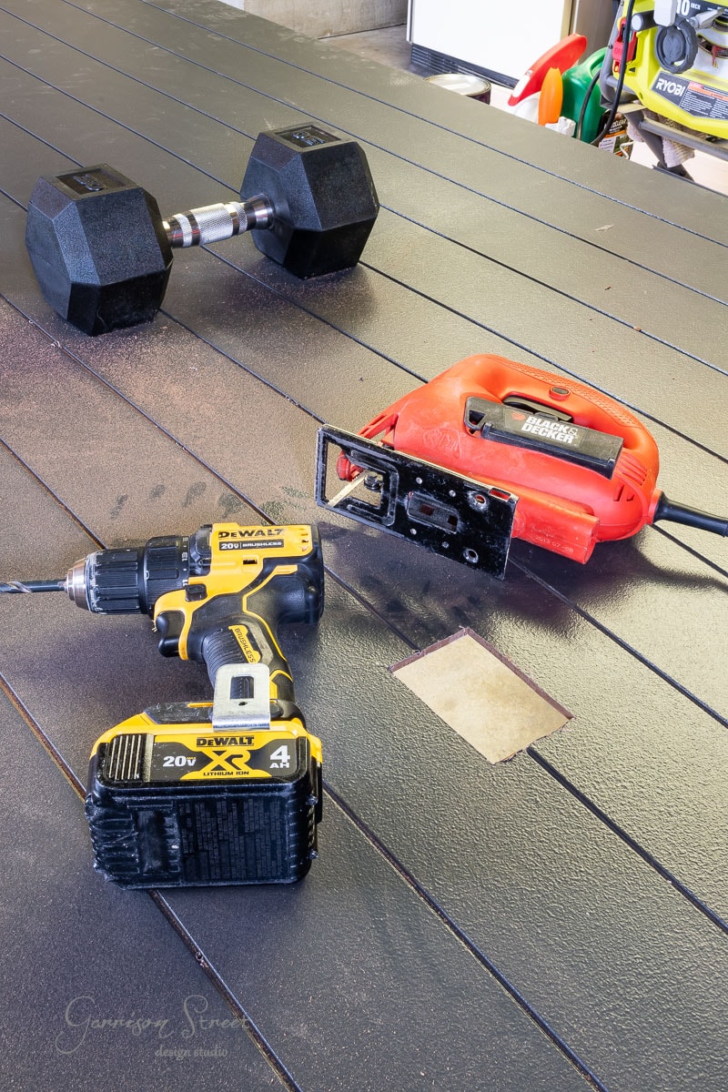 A workbench features a black dumbbell, a yellow cordless power drill, and a red jigsaw. The surface is sprinkled with wood dust and shows a small cut-out section. In the background, various colorful tools and equipment complete the scene.