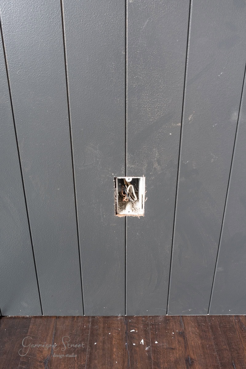 A dark wooden wall panel with vertical grooves features a square opening for an electrical outlet with exposed wires. The floor showcases a polished wooden surface.