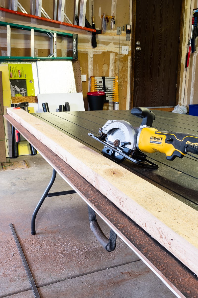 A circular saw rests on a wooden workbench in a garage. Shelves with various tools and materials sit in the background, including a ladder, containers, and wood pieces. This well-lit setting showcases a tidy and organized workspace.