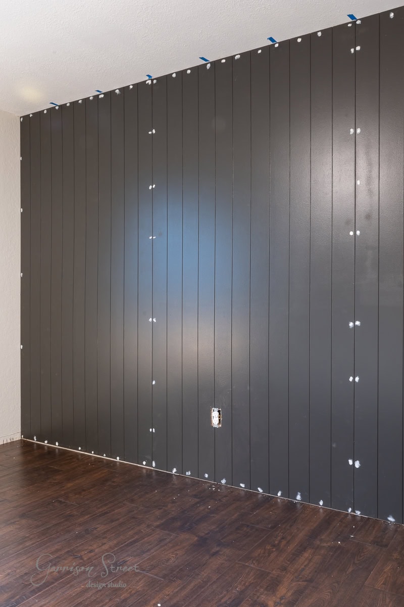 A modern DIY shiplap accent wall features dark vertical paneling with visible screws, complemented by a floor of dark wood planks. A visible electrical outlet graces the lower wall, while blue tape runs along the top edge for precision.