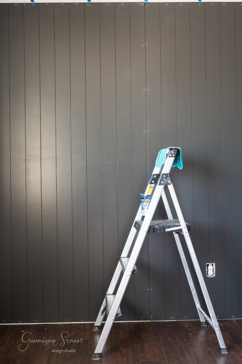 A DIY shiplap accent wall painted black serves as the backdrop, with a silver ladder positioned in front. A blue cloth and a small container rest on the ladder's step, contrasting elegantly against the dark wood floor.